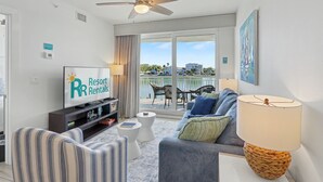 A living room with a television displaying 'Resort Rentals,' a ceiling fan, a sofa, a coffee table, and a view of a pool and buildings.