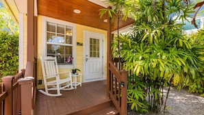 Une véranda en bois avec une chaise berçante et une petite table, une porte blanche et de nombreuses plantes vertes luxuriantes.