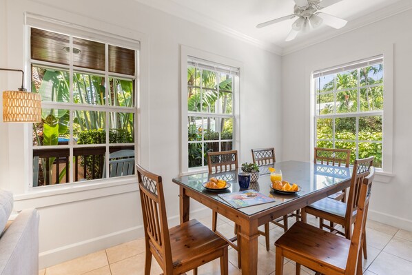 A dining area with a wooden table and chairs, large windows with views of greenery, and a ceiling fan.