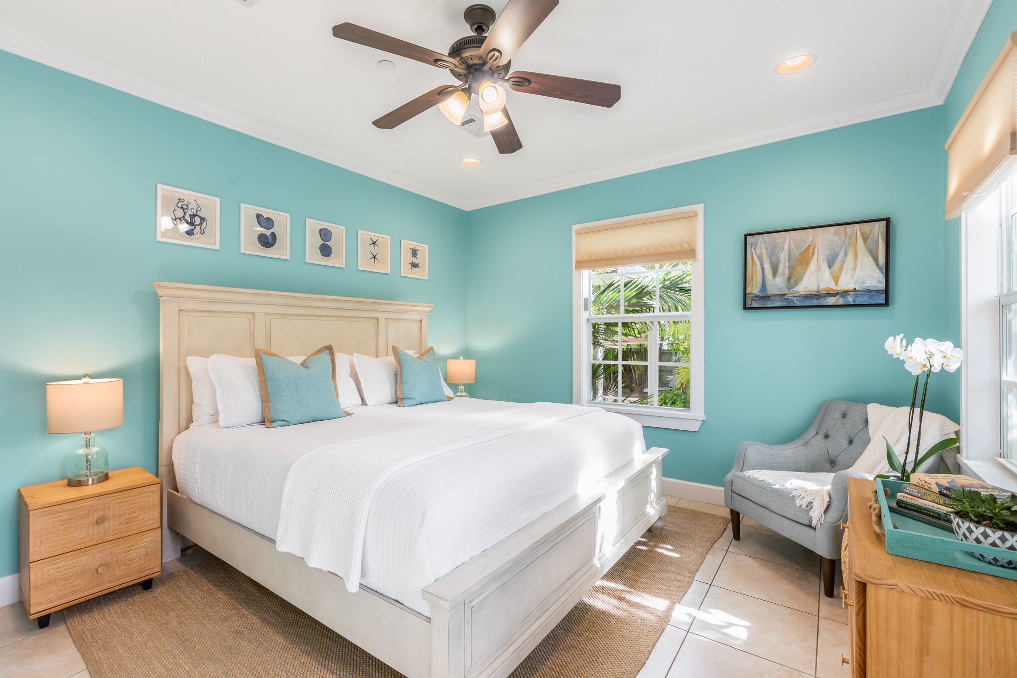 A bedroom with a large bed, a ceiling fan, a nightstand, a chair, and a window with a view of greenery.
