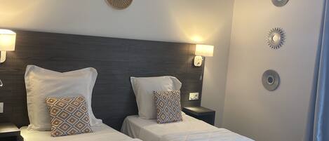 Een hotelkamer met twee enkele bedden, een hoofdbord van donker hout, bedlampjes, een wandspiegel en een ronde lamp.