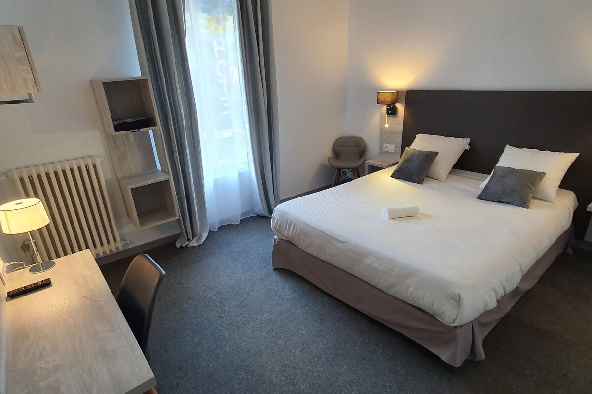 A hotel room with a bed, bedside tables, a desk, a chair, a lamp, and a window with curtains.
