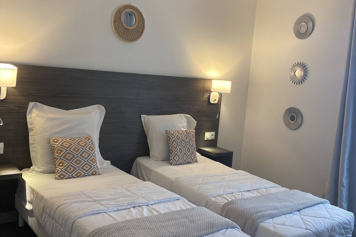 A hotel room with two beds, a dark headboard, wall sconces, a round mirror, and a small table with a lamp.