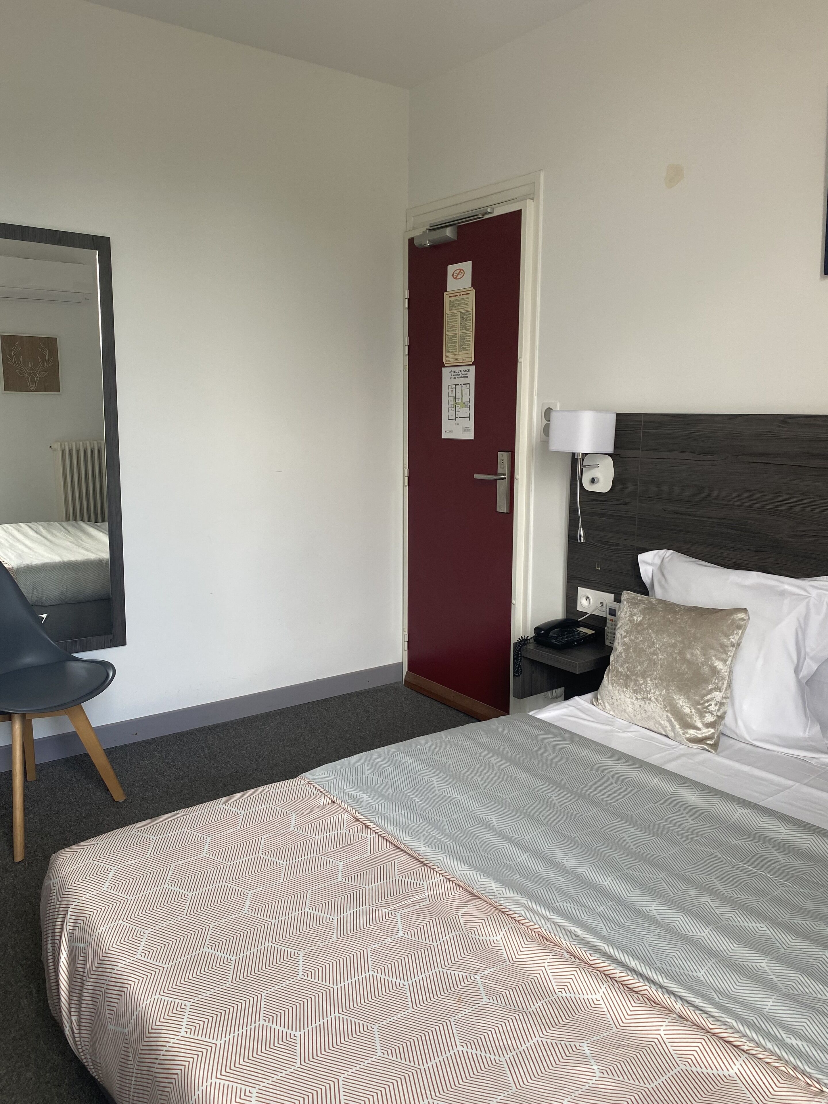Hotelkamer met een bed, stoel, nachtkastje, lamp en een deur met prikbord.