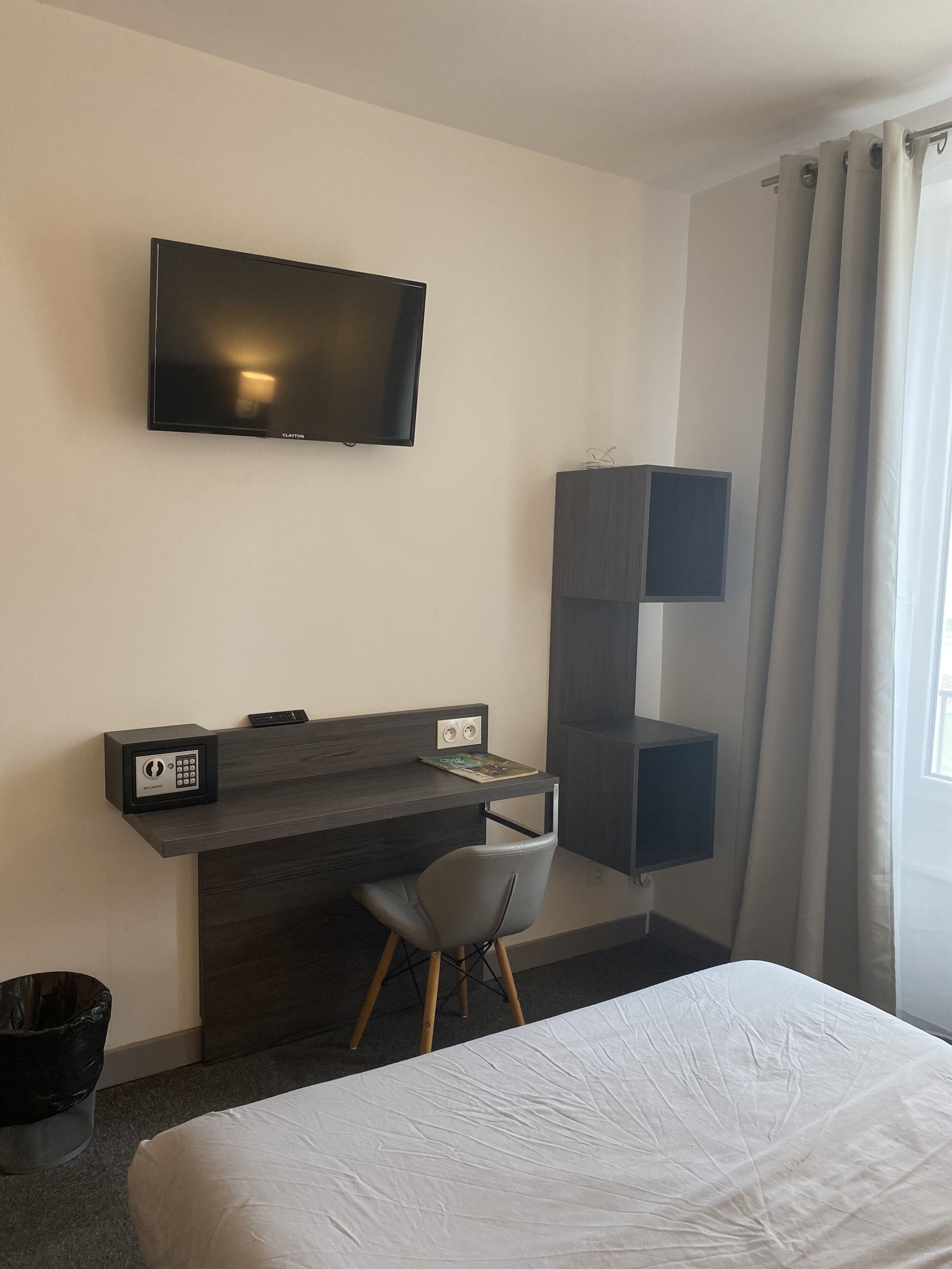 A hotel room with a bed, a desk with a chair, a TV mounted on the wall, and a window with curtains.
