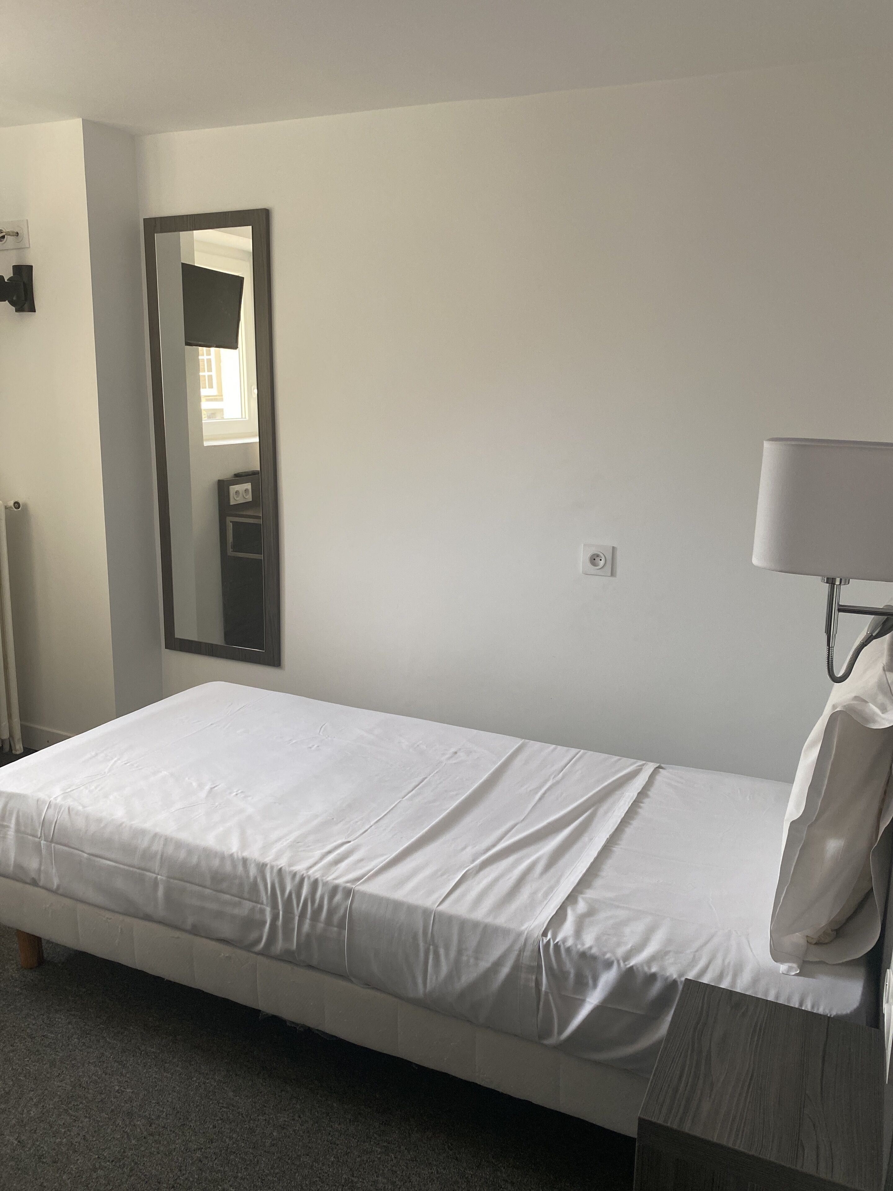 Habitación de hotel sencilla y limpia con una cama individual, una lámpara de noche, un espejo y un radiador.