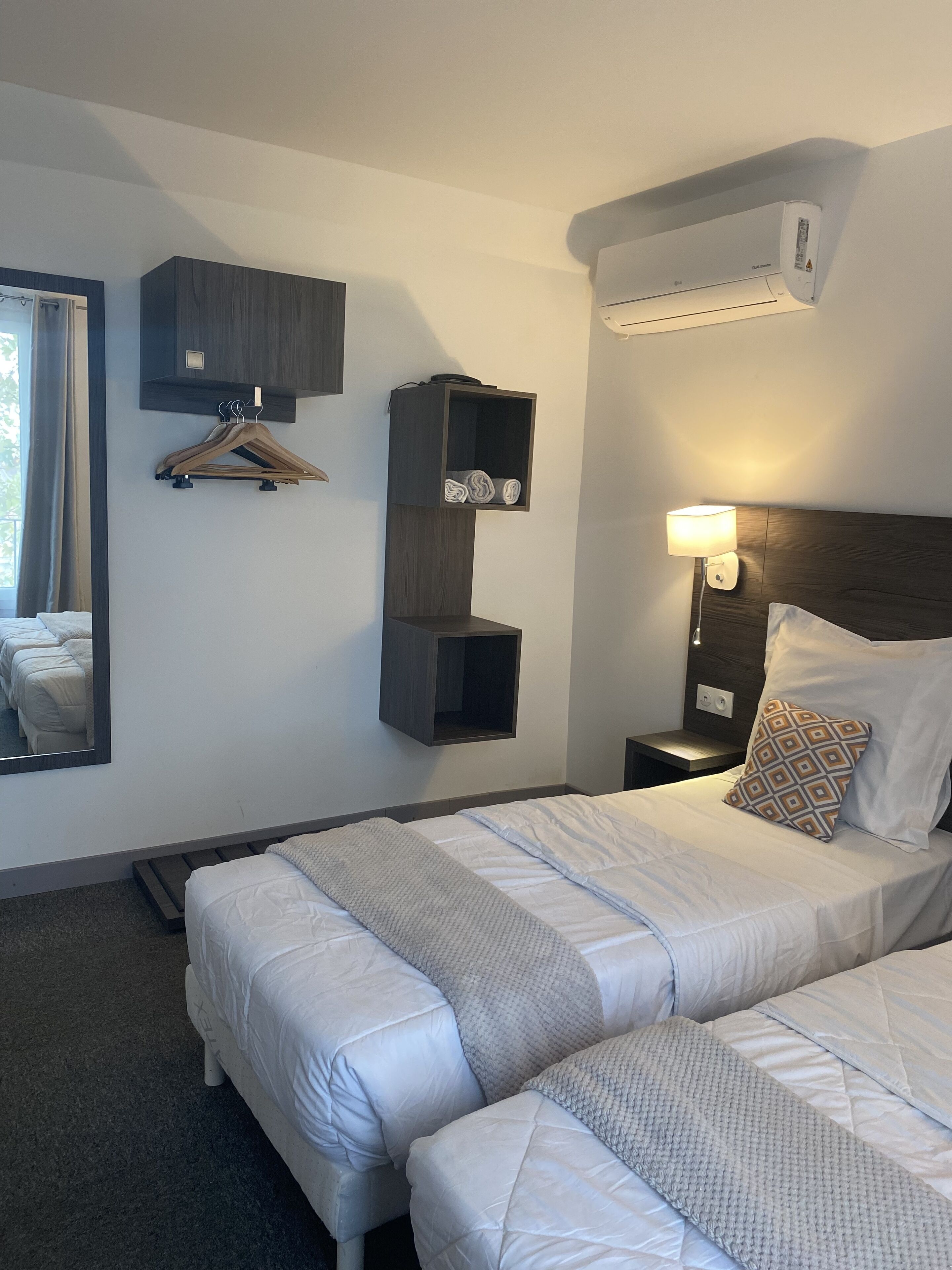 Habitación de hotel con dos camas, un armario, un espejo, una mesita de noche con lámpara y un equipo de aire acondicionado.