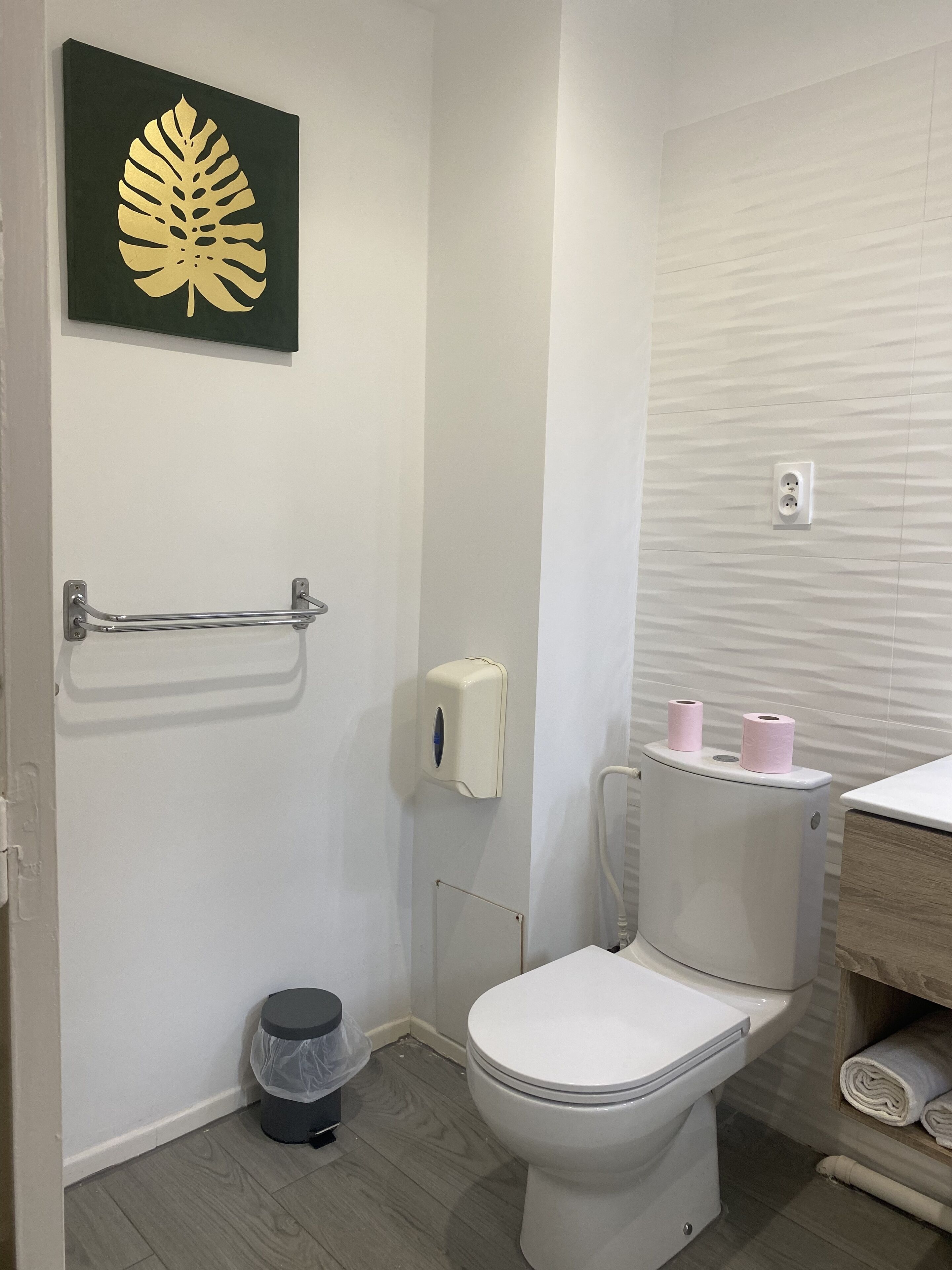 A bathroom with a toilet, a trash can, a towel rack, a soap dispenser, and a wall-mounted outlet.