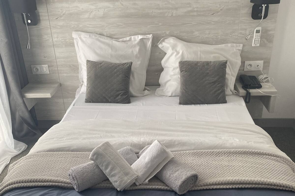 A neatly made bed with white and gray pillows, a white blanket, and rolled towels. There is a bedside table with a phone and a lamp.