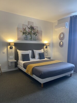 A modern bedroom with a large bed, two bedside tables, wall-mounted lamps, and a decorative wall piece above the bed. - Hôtel l'Alsace (Narbonne)
