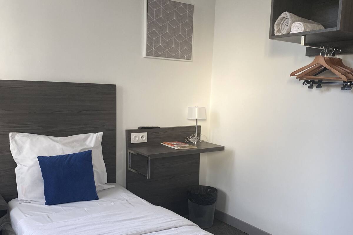 A neatly made bed with white and blue pillows, a bedside table with a lamp, a small trash bin, and a wall-mounted shelf with a towel and hangers.