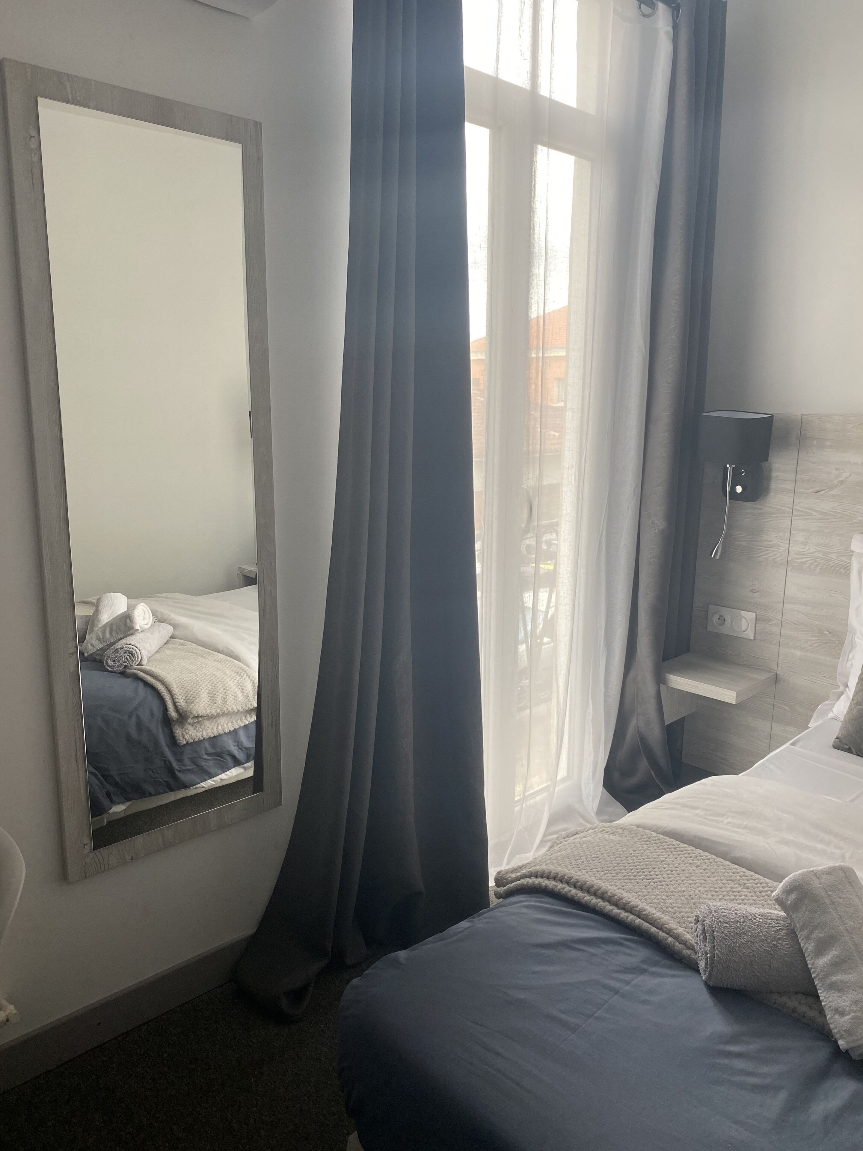 A bedroom with a large mirror, a bed with a blue and white duvet, a bedside table, and a wall-mounted lamp.