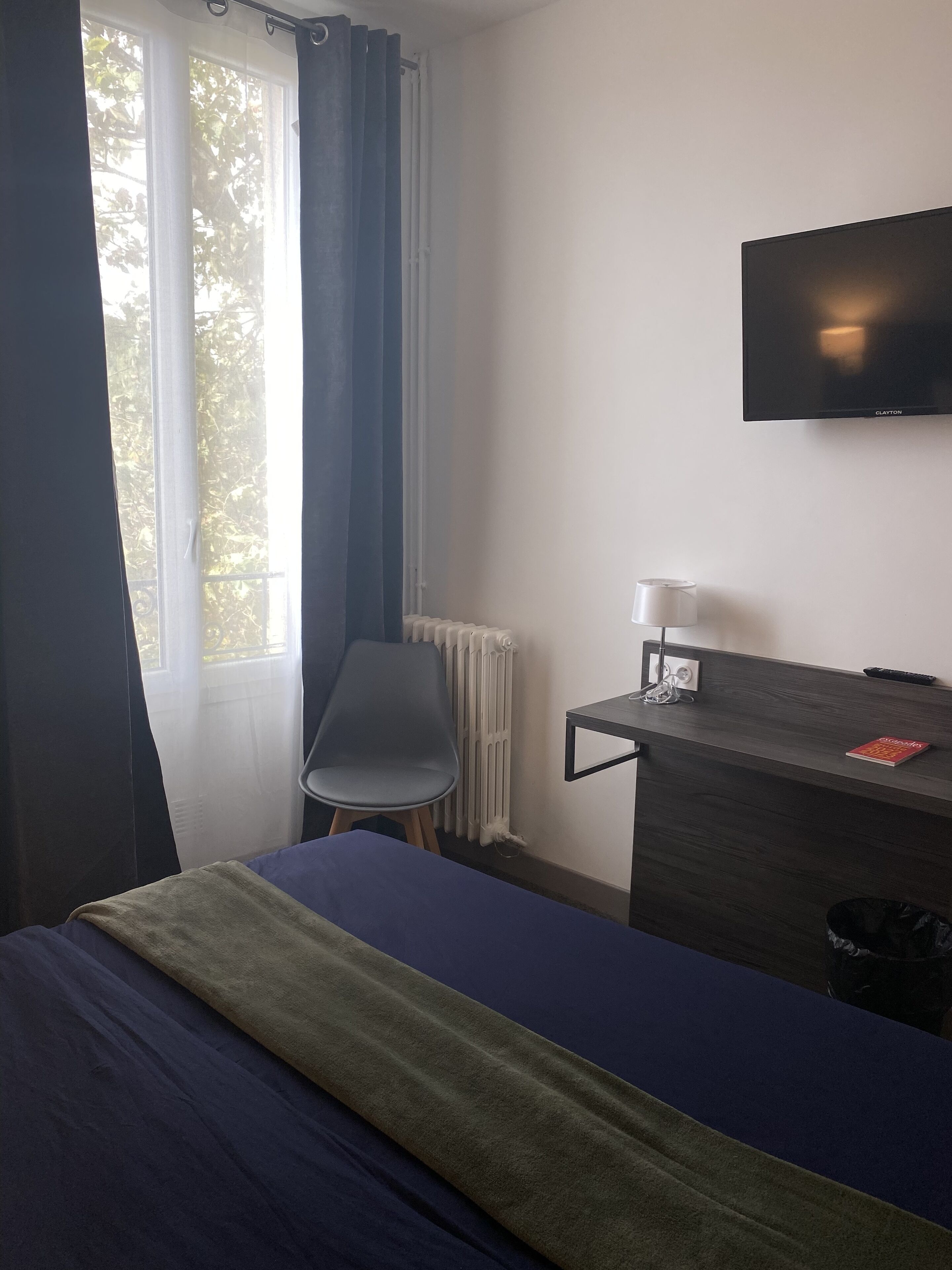 A hotel room with a bed, a desk with a lamp, a chair, a radiator, and a TV on the wall.