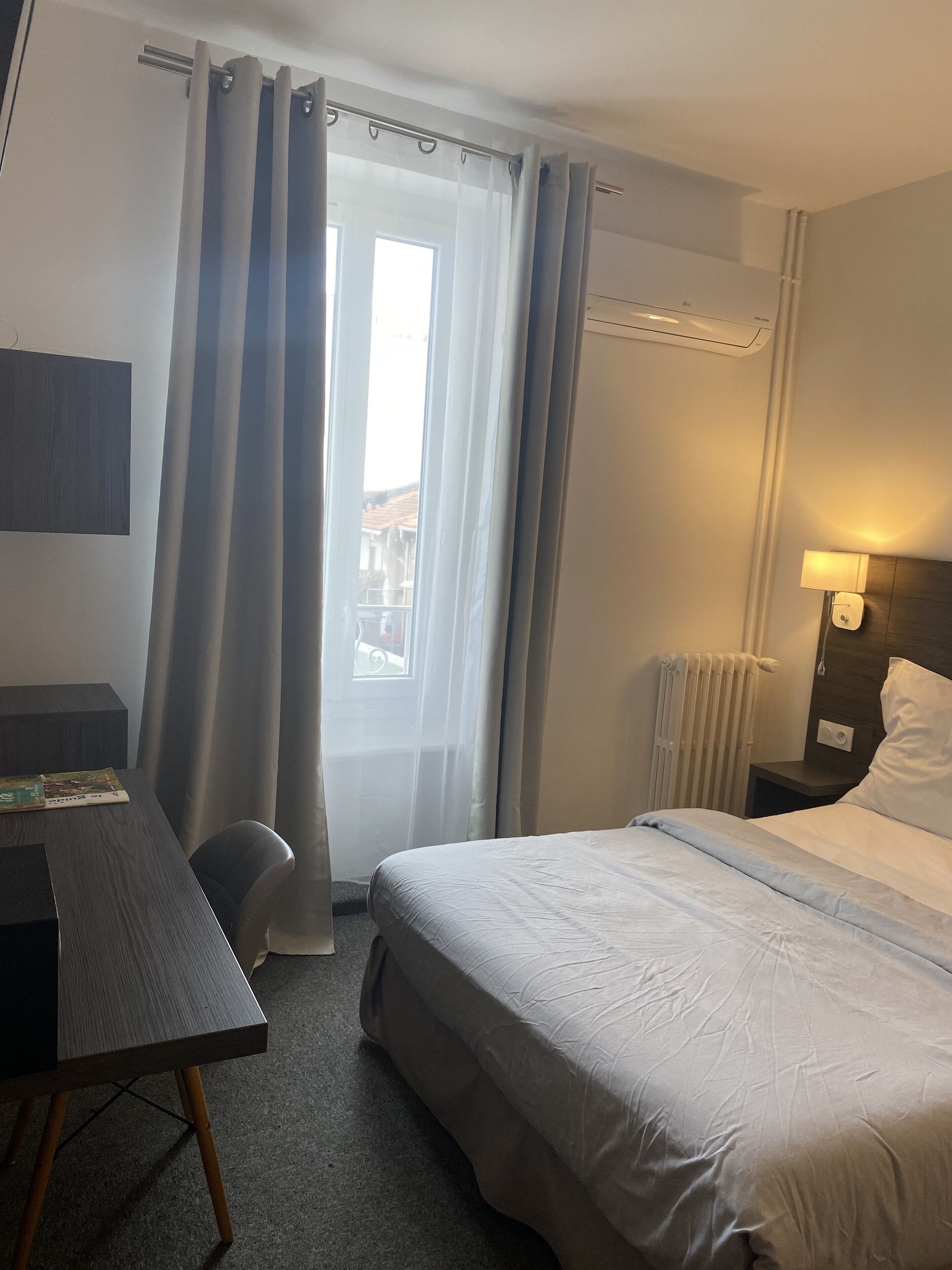 Quarto de hotel com cama, escrivaninha, cadeira, mesa de cabeceira com abajur e janela com cortinas.