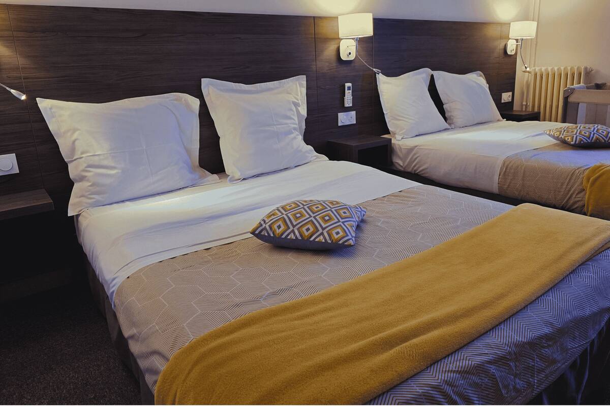 A hotel room with two beds, each with white linens and patterned bedspreads. There are wall-mounted lamps and a thermostat on the wall.