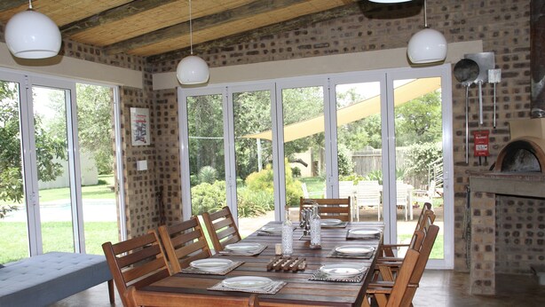 A dining area with a large wooden table set for six, surrounded by wooden chairs, with a brick wall and a stone fireplace.