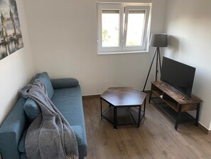 En stue med en blå sofa, et lille træbord, en gulvlampe og et fjernsyn på et træstel.