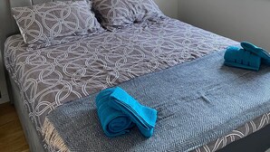 Una cama bien tendida con ropa de cama estampada y toallas enrolladas, un estante de madera con adornos, y una ventana con luz natural.
