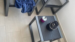 Un balcón con dos sillas de metal gris, una manta azul, una mesita con un recipiente morado y una cesta negra.