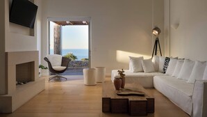 Un salon moderne avec un canapé blanc, un téléviseur à écran plat fixé au mur, une cheminée et une vue sur l’océan par une porte ouverte.