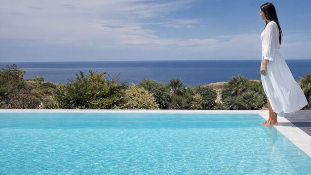 Une femme vêtue d’une robe blanche se tient près d’une piscine, avec vue sur l’océan.