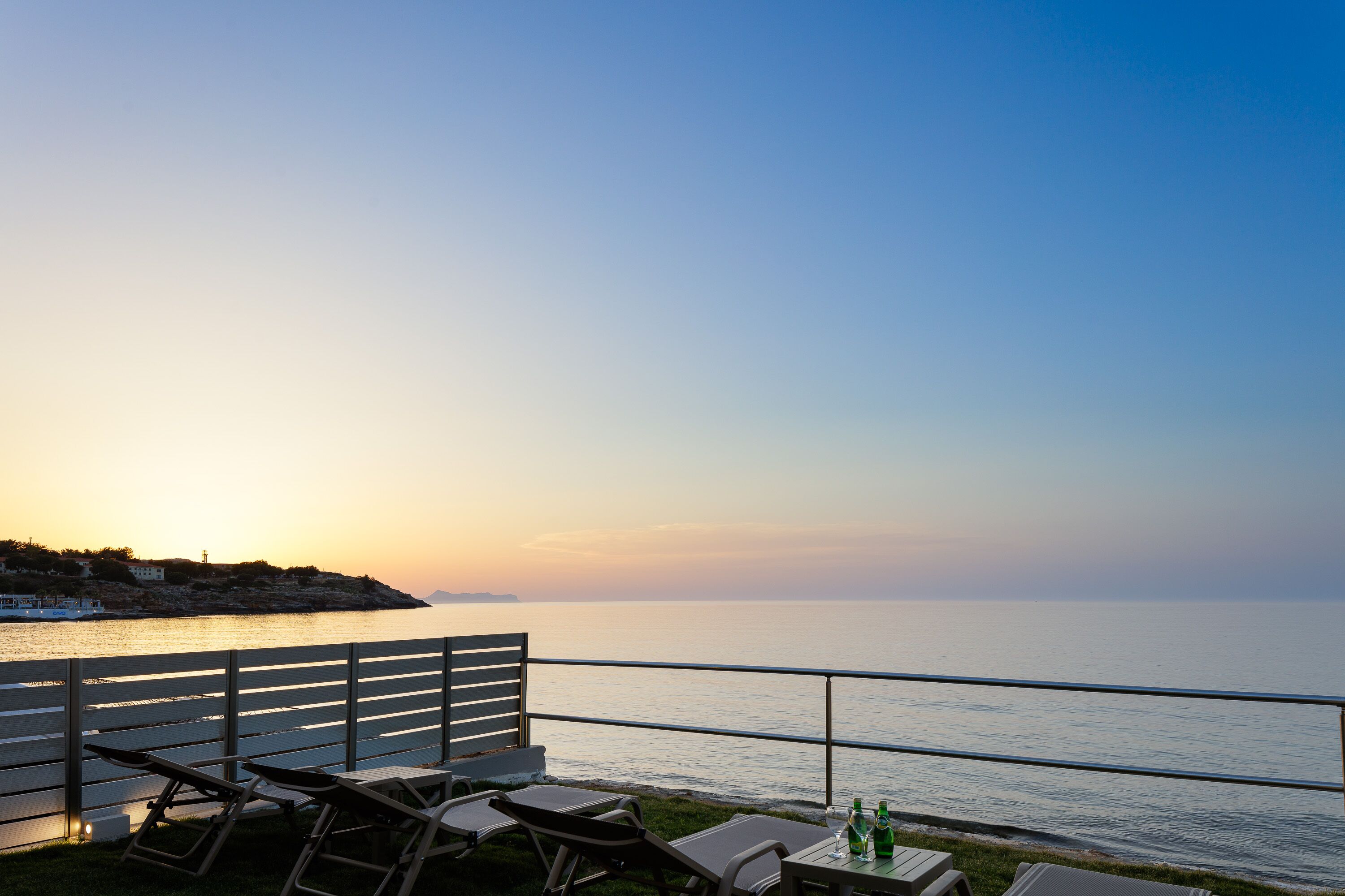 Une terrasse avec des fauteuils et une table, d’où l’on peut admirer une mer calme et un coucher de soleil.