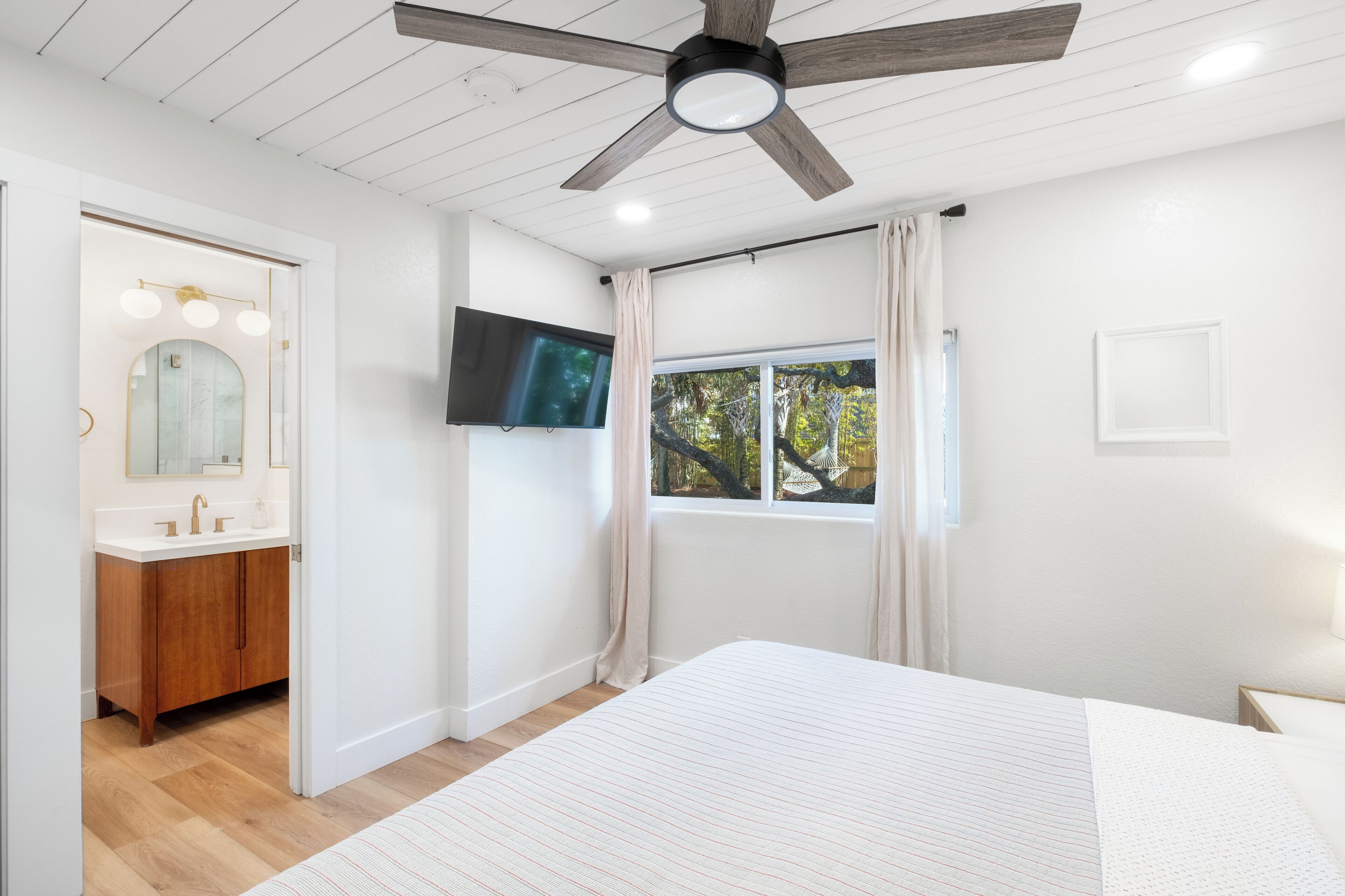 A bedroom with a bed, a ceiling fan, a TV, a window with curtains, and a bathroom with a vanity and mirror.