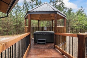 A wooden gazebo with a hot tub, surrounded by a wooden deck and metal railings, set in a forested area. - Geauxin' All Inn -Gameroom, Bunks, Spacious Covered Deck, Paved Rd, Hot Tub (Broken Bow)