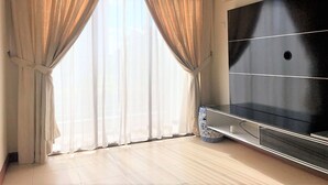 Una habitación con un televisor de pantalla grande, una ventana con cortinas translúcidas y piso de baldosas.