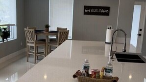 Une cuisine moderne dotée d’un grand îlot central, d’un coin repas avec des chaises, d’une fenêtre et d’une inscription murale qui dit : « L’amour commence à la maison ».