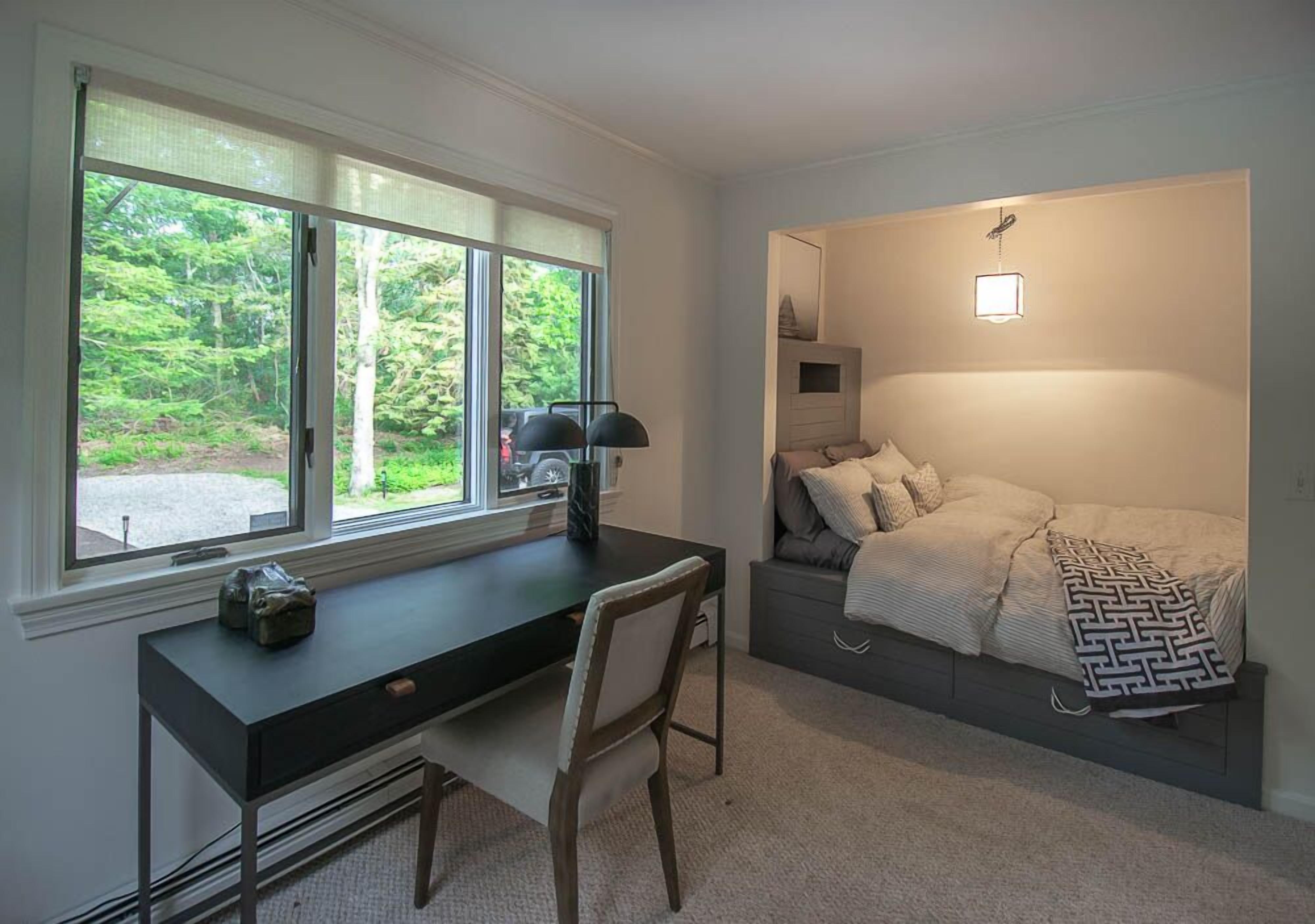 Une chambre à coucher avec un lit, un bureau et une chaise. Il y a une fenêtre qui donne sur des arbres.