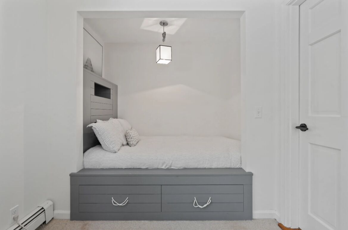 Une chambre à coucher avec un lit, une commode grise dotée de tiroirs, un luminaire suspendu et une porte blanche.