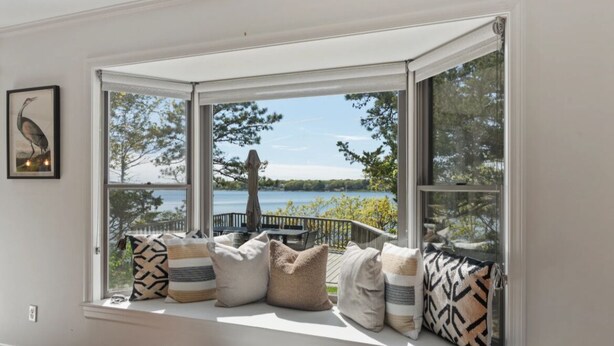 A bay window with a view of a lake and trees, a wooden deck, and several pillows on the window seat. - 2 Acre Waterfront Home Overlooking Waquoit Bay with Spa and Swim Dock  (Falmouth)