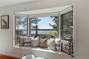 A bay window with a view of a lake and trees, a wooden deck, and several pillows on the window seat. - 2 Acre Waterfront Home Overlooking Waquoit Bay with Spa and Swim Dock  (Falmouth)