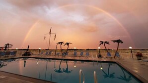 Zwembad met ligbedden en palmbomen, onder een lucht met een regenboog.