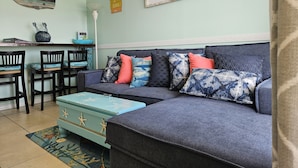 Ein gemütliches Wohnzimmer mit einem grauen Sofa, gemusterten Kissen, einer blauen Kommode und einem 'WELCOME'-Schild an der Wand.