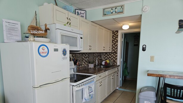 A compact kitchen with white appliances, a tiled backsplash, and a small dining area with a chair and table. - Beachfront with Ocean Views right from your balcony (Wildwood Crest)