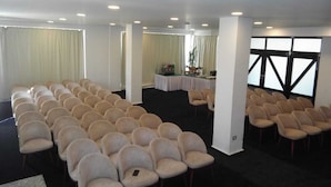 A conference room with rows of beige chairs, a stage with a table, and large windows with blinds.
