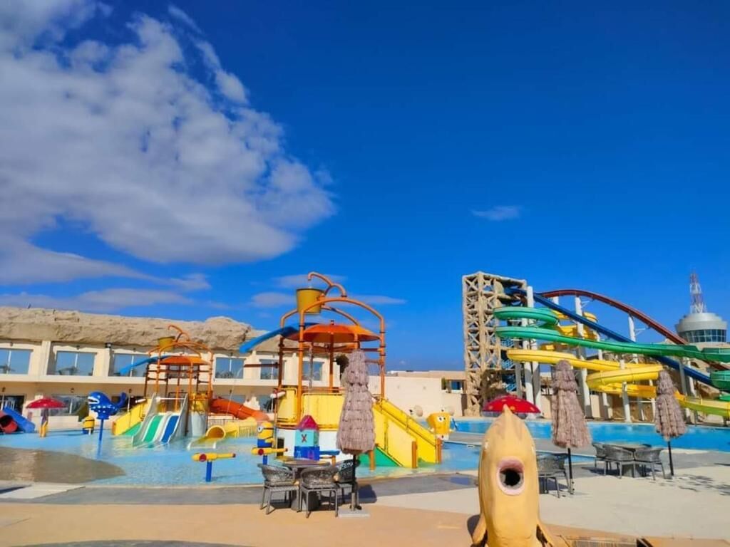 A water park with slides and a wave pool, a lifeguard tower, and a poolside area with umbrellas and lounge chairs.
