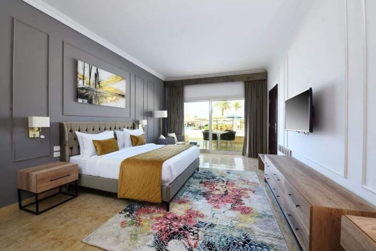 A modern hotel room with a large bed, a wooden dresser, a flat-screen TV, and a view of the outdoor area.