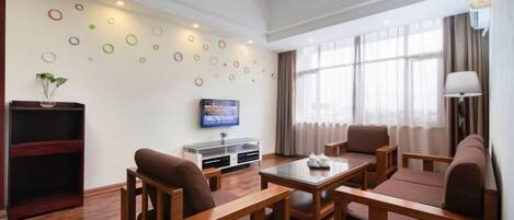 A hotel room with a wooden floor, a TV, a sofa, a coffee table, and a bookshelf.