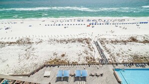 Vista aérea de una playa con filas de tumbonas y sombrillas, una piscina y una torre de salvavidas.