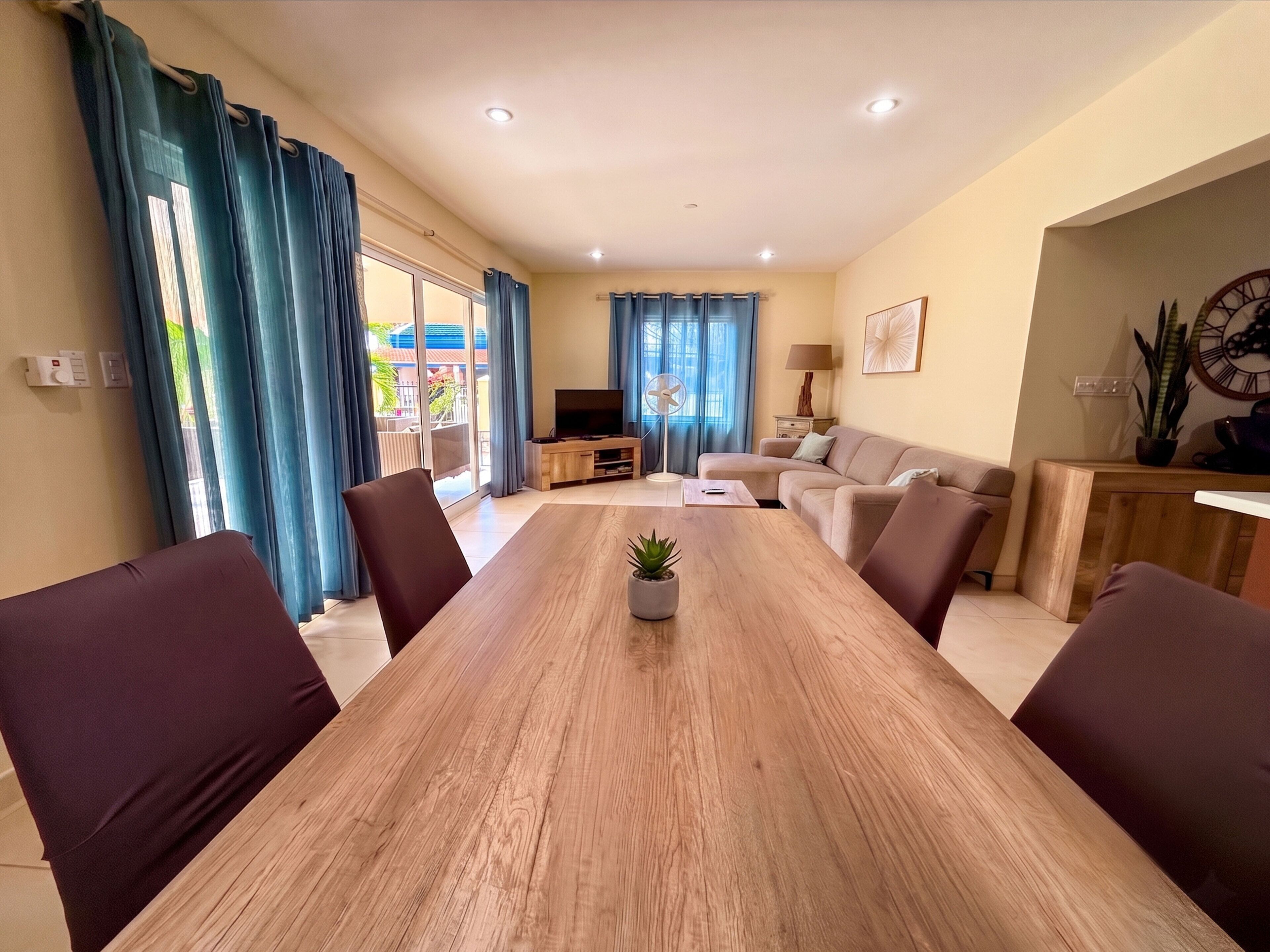 A modern living room with a wooden dining table, sofas, a TV, and a balcony with blue curtains.