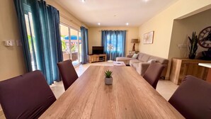 Un salon moderne avec une table à manger en bois, des canapés, un téléviseur et un balcon agrémenté de rideaux bleus.