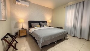 A bedroom with a bed, two bedside tables, a lamp, a hanging light, a window with curtains, and an air conditioning unit.