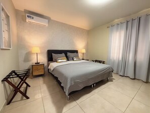 A bedroom with a bed, two bedside tables, a lamp, a hanging light, a window with curtains, and an air conditioning unit. - Bonairechoice oceanfront apartment (Kralendijk)