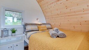 Ett sovrum med ett träpanelat tak, en säng med ett gult, vadderat överkast, ett sängbord med en klocka och en växt, och ett fönster med utsikt över grönskan.