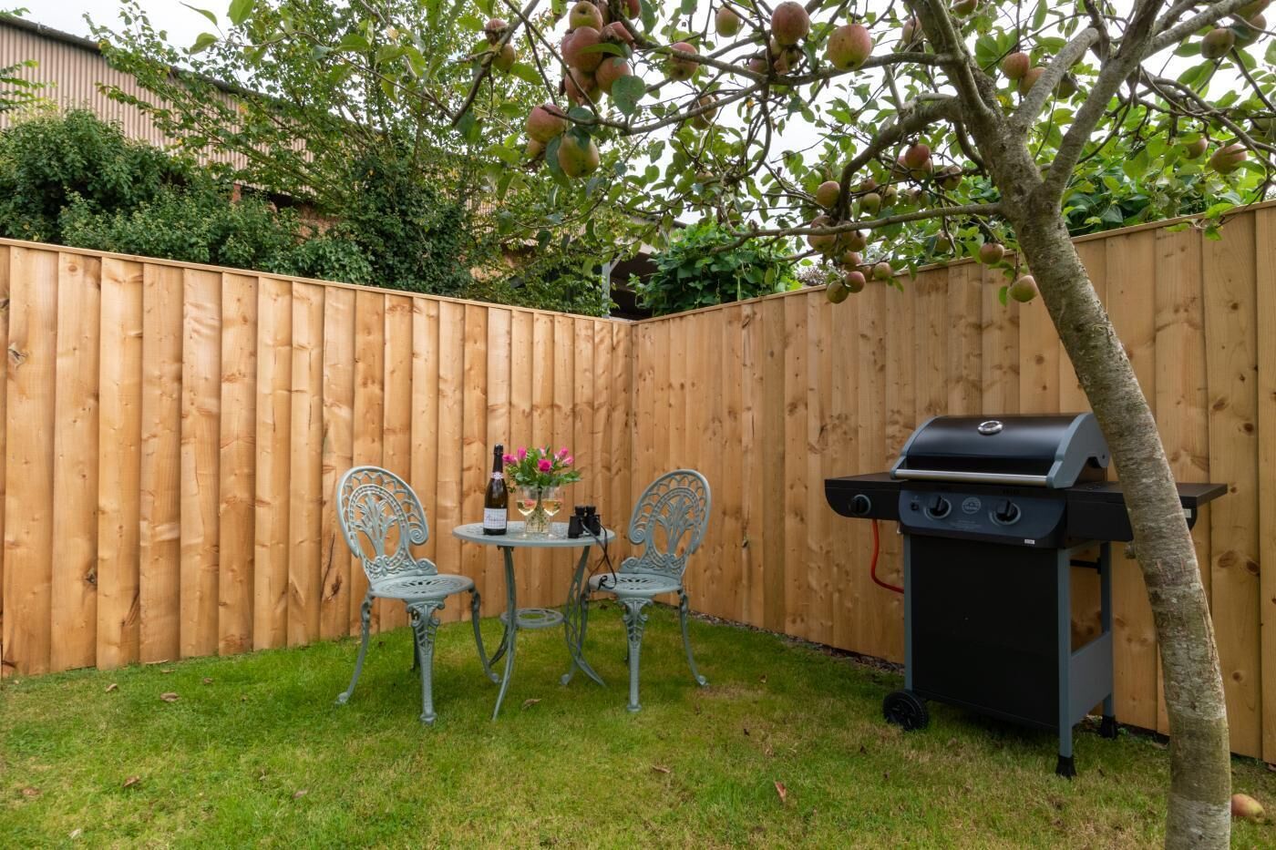 A backyard with a wooden fence, two metal chairs, a small round table with a bottle and flowers, and a barbecue grill.