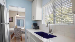 A kitchen with a blue sink, white cabinets, a coffee maker, and a dining area with patterned chairs.