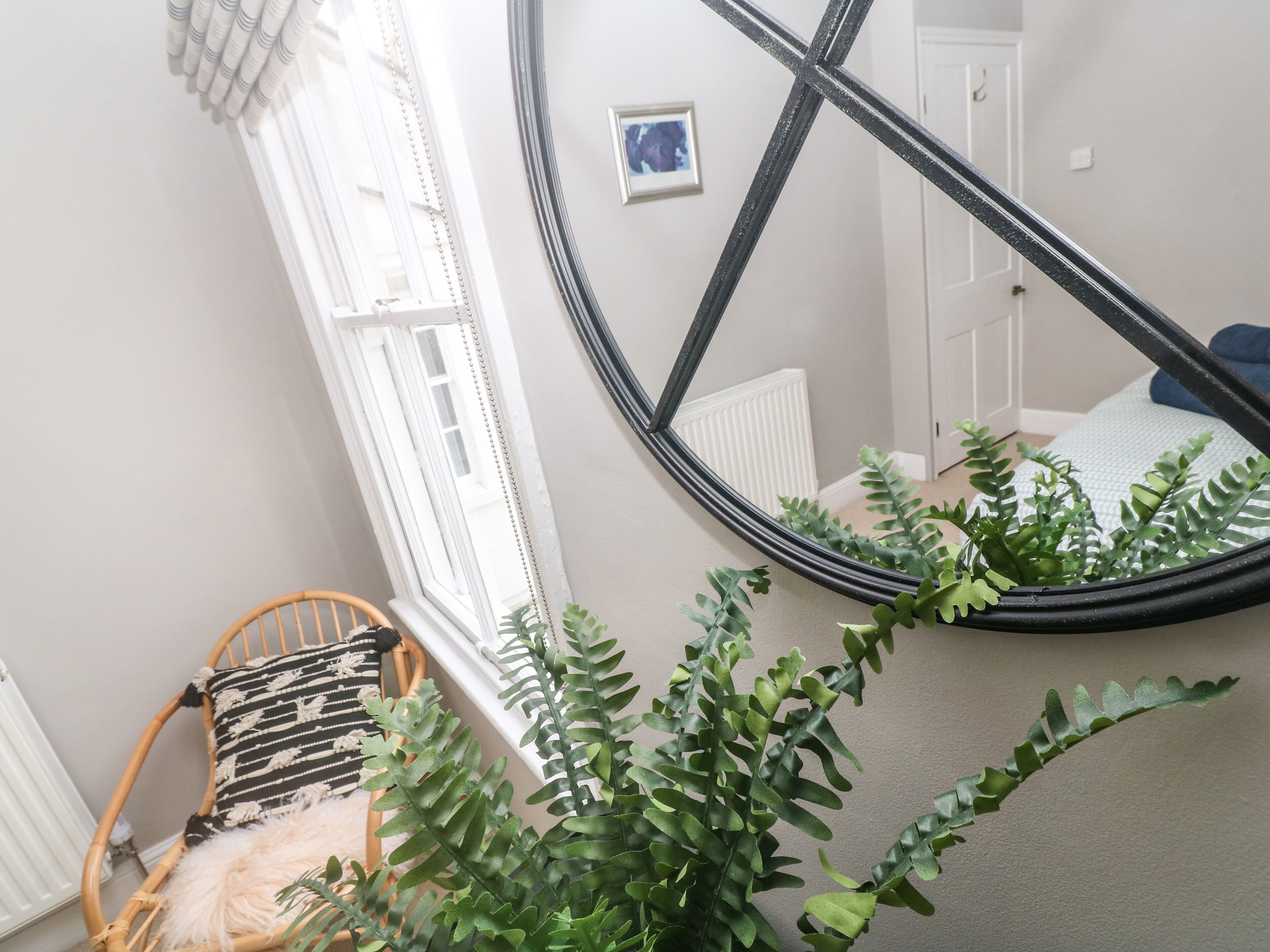 Une pièce avec un grand miroir qui reflète un lit, une fenêtre avec des stores, une chaise en osier et une plante en pot.